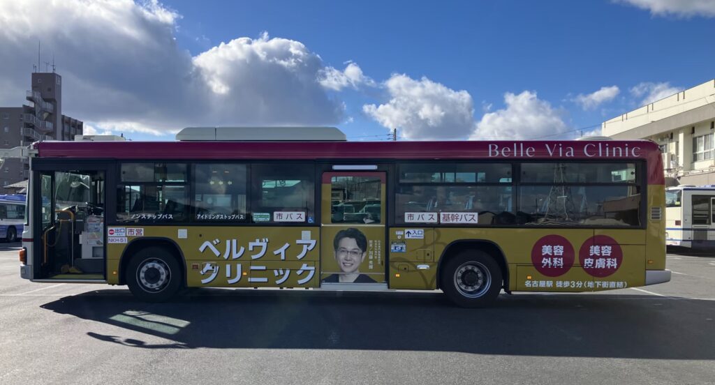名古屋駅周辺を走行するベルヴィアクリニックのラッピングバス側面。形成外科専門医である加藤 秀輝院長の肖像と、美容外科・美容皮膚科の診療案内、名古屋駅徒歩3分（地下街直結）の文字がデザインされています。